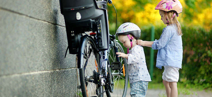 Two small children wearing cycle helmets are standing next to an adult’s bicycle. The children are wearing summer clothes, and verdant plants can be seen in the background.