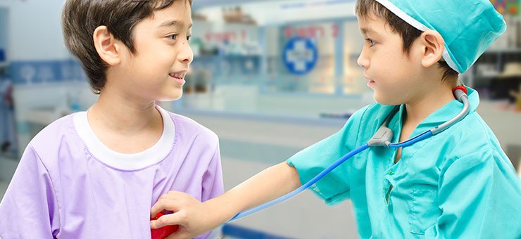 Two small children are playing doctor and patient. One of them is wearing green surgical scrubs and a hat, the other mauve surgical scrubs. The child wearing green is listening to the other child’s heart through a stethoscope.