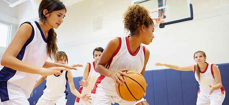 خمس فتيات مراهقات يلعبن كرة السلة فريق ذو قمصان بيضاء وحمراء، والفريق الآخر بقمصان بيضاء وزرقاء.