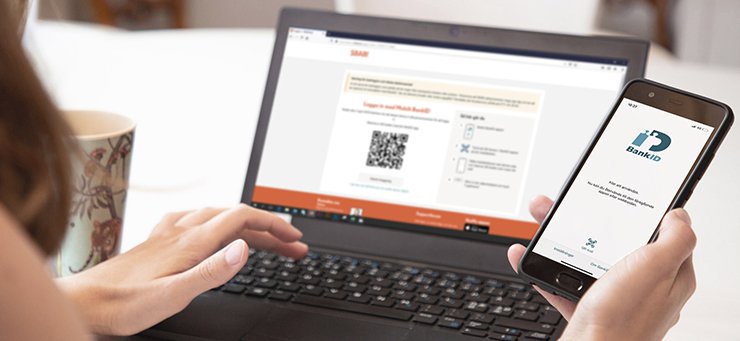En kvinna sitter vid en laptop, på skärmen syns en online banksida. I ena handen håller hon en mobiltelefon med appen mobilt bank-id öppen.