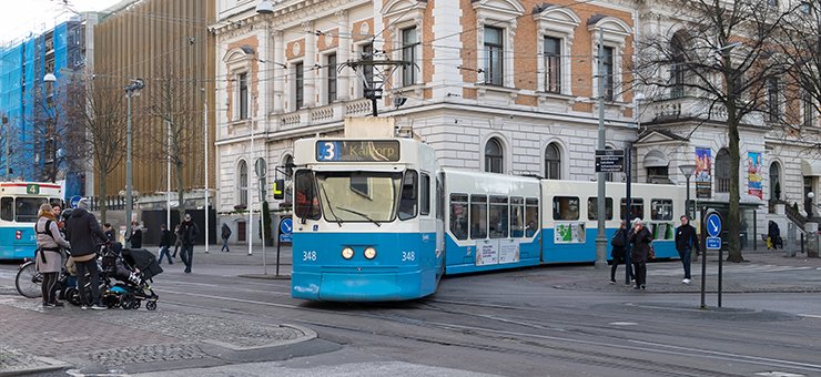 Street view from Gothenburg. A blue and white tram is turning towards the camera while another is disappearing from view on the left. A number of people can be seen around the trams.