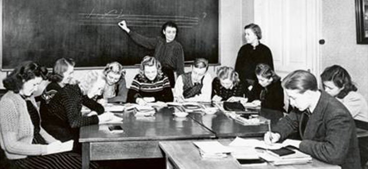 Dans une salle de classe, des adultes sont assis autour de différentes tables dans le cadre d’une formation pour adultes au début du 20ème siècle.