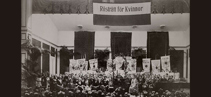 An assembly hall in the early 20th century with women and various placards and a banner reading “Voting rights for women”.