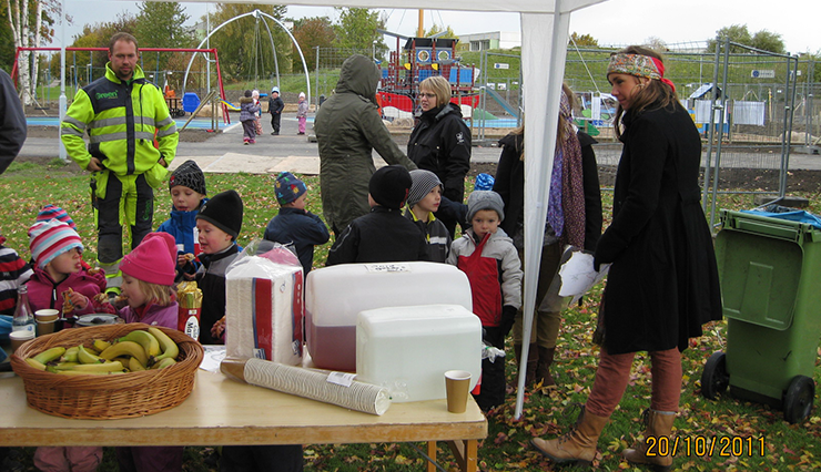 En grupp små barn står under ett tält. Framför barnen står ett bord uppdukat med saft och frukt. I bakgrunden finns en lekplats.