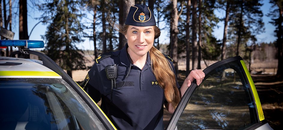 Une policière à côté d’une voiture de police avec des arbres en arrière-plan.